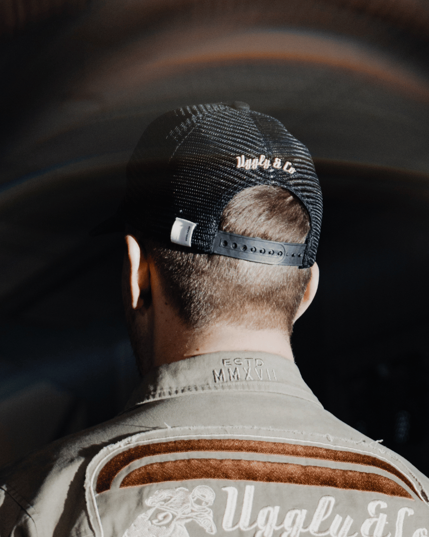 Rear view of a model wearing an all-black trucker hat with a vintage-inspired flat track racing logo in yellow and blue on the front at night