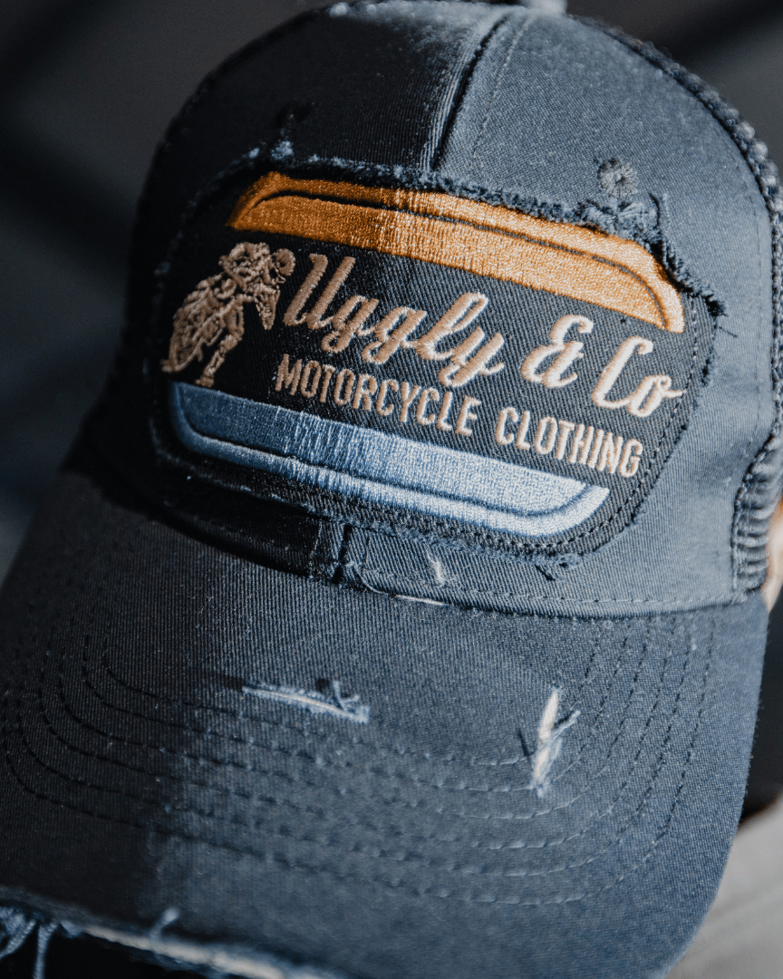 An all-black trucker hat with a vintage-inspired flat track racing logo in yellow and blue on the front with a dark blurred background