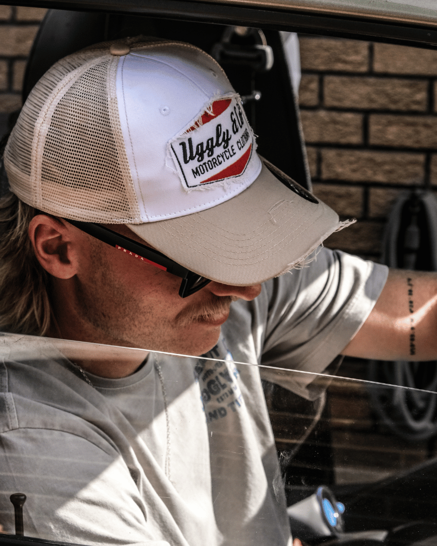 Side view of a model driving a car while wearing a beige and white Uggly & Co motorcycle clothing trucker hat with a mesh back and an embroidered logo on the front