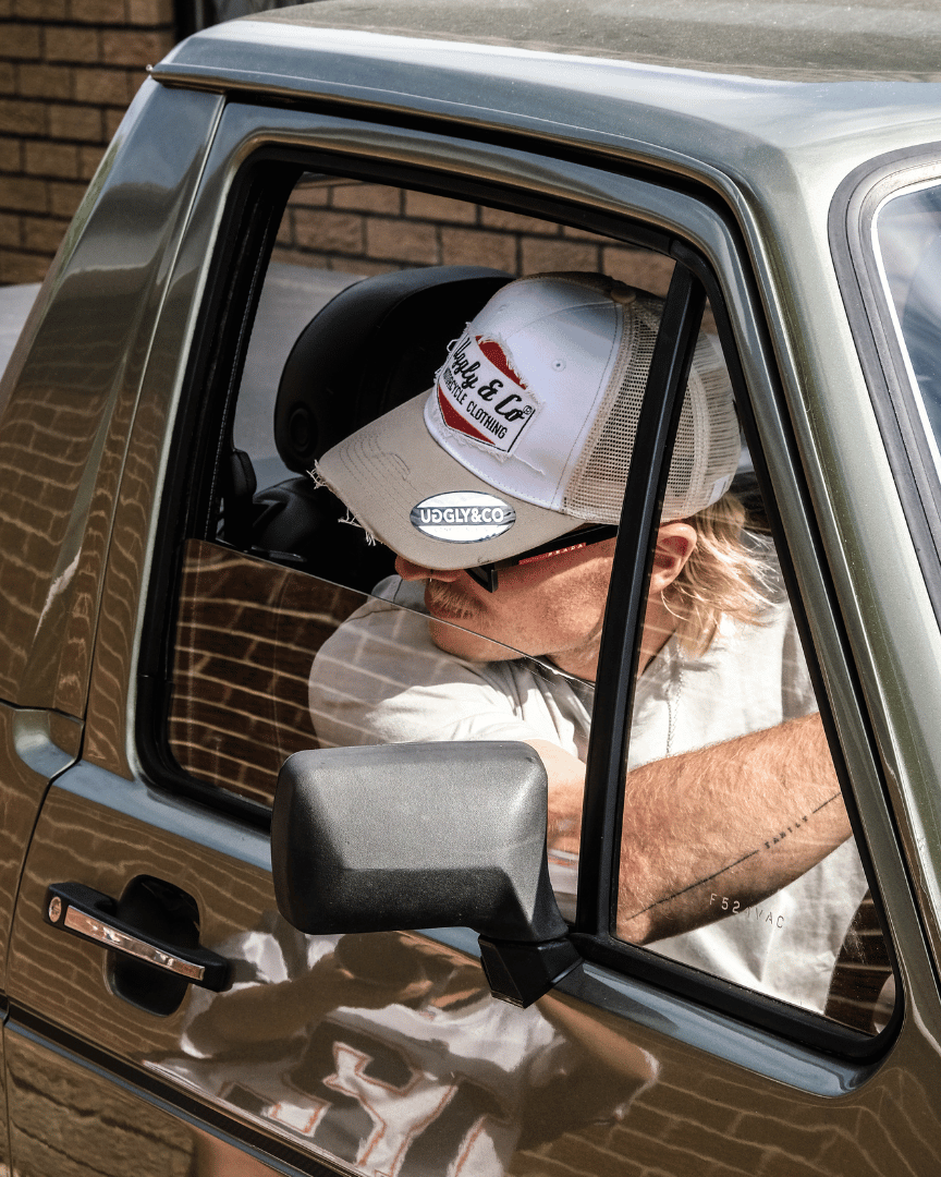 A model driving a car while wearing a beige and white Uggly & Co motorcycle clothing trucker hat with a mesh back and an embroidered logo on the front