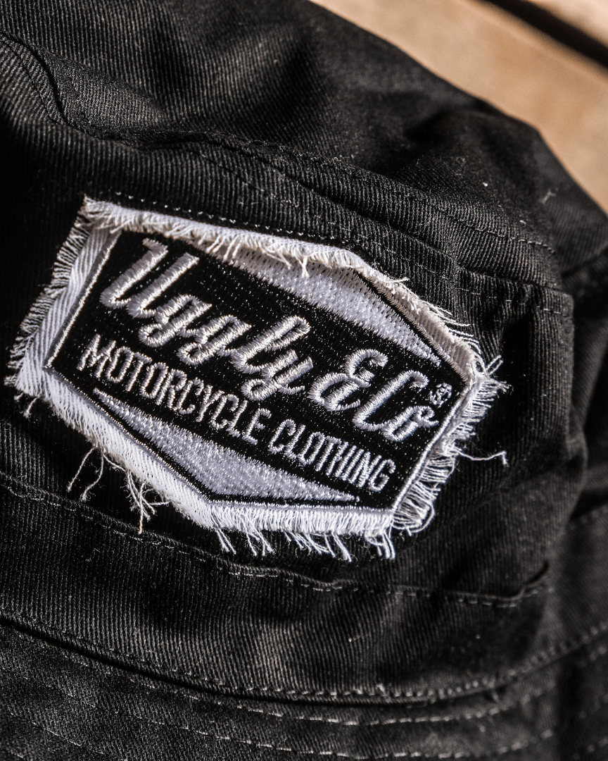 Close up of a black bucket hat with a distressed diamond Uggly&Co logo displayed on a wooden background.