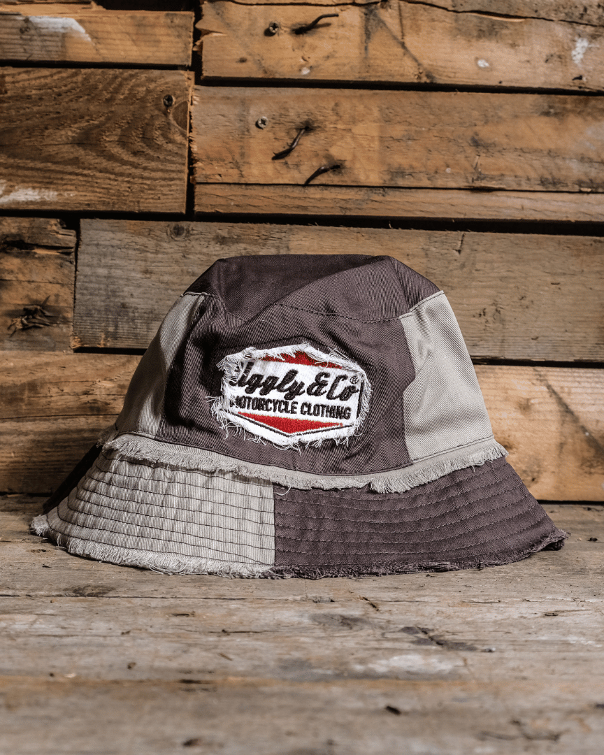 A beige and brown patchwork bucket hat with frayed edges, featuring a diamond Uggly&Co logo, displayed on a wooden surface.