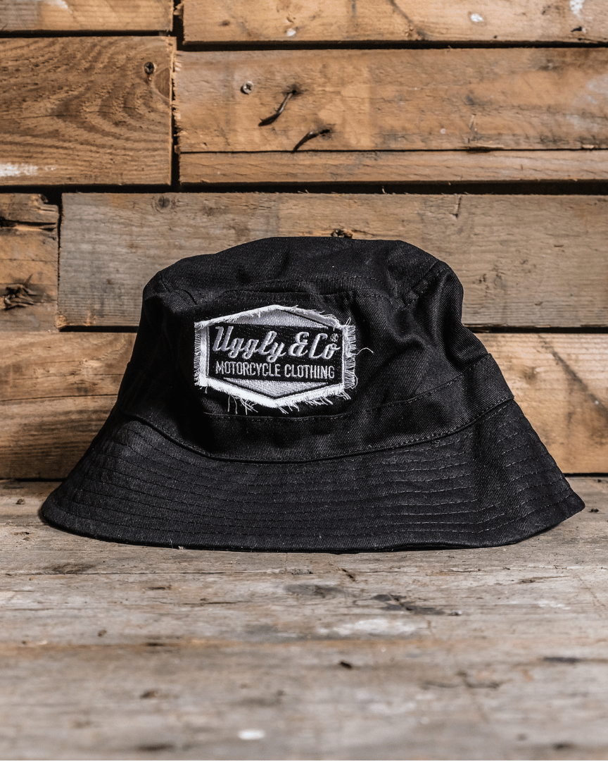 A black bucket hat with a distressed diamond Uggly&Co logo displayed on a wooden background.