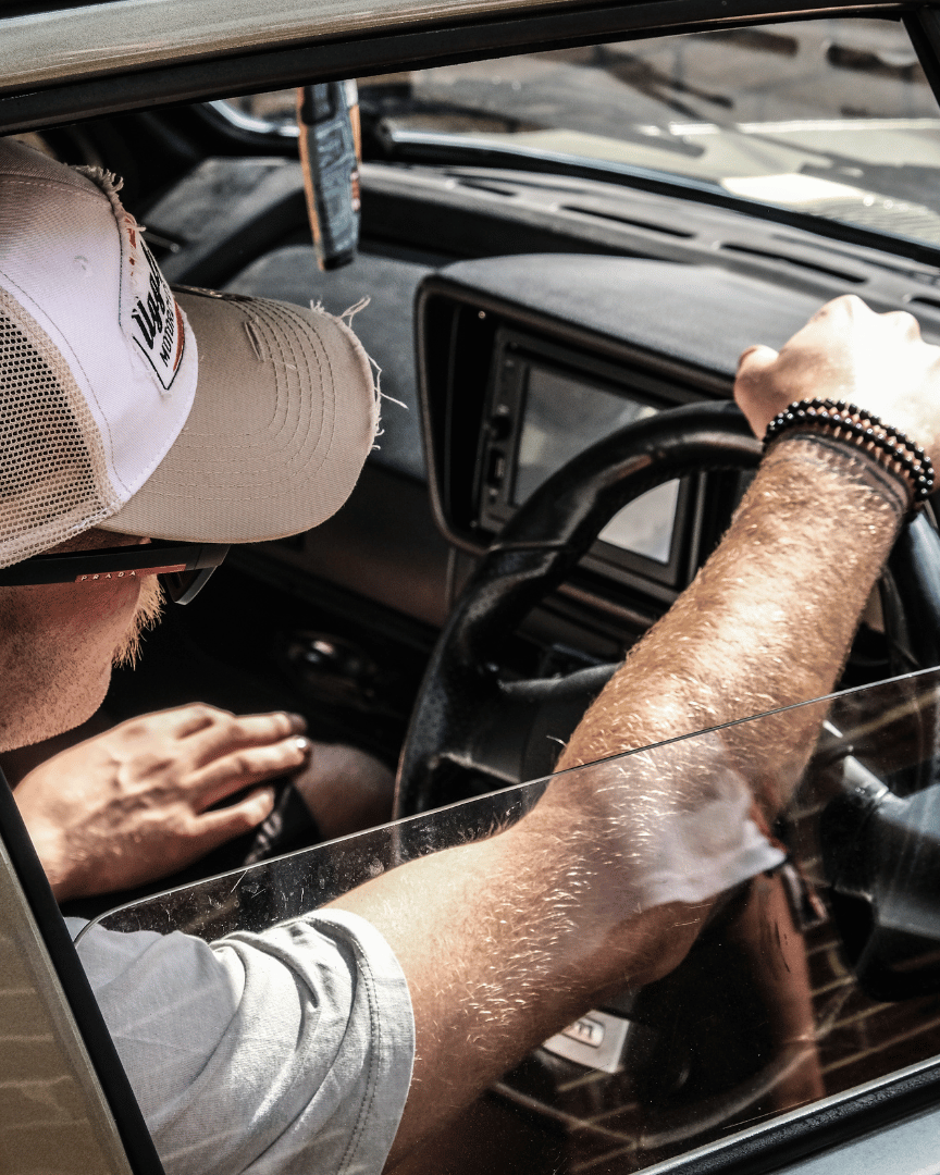 Diagonal view of a model driving a car while wearing a beige and white Uggly & Co motorcycle clothing trucker hat with a mesh back and an embroidered logo on the front