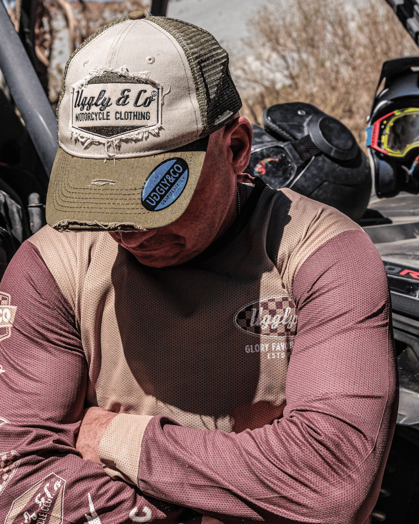 A motocross rider standing in the desert in a brown Uggly&Co MX shirt wearing a cap