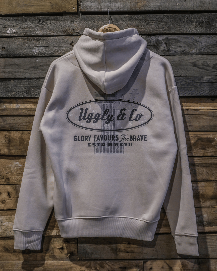 A gray hoodie with the brand name 'Uggly&Co' and the text 'GLORY FAVOURS THE BRAVE' printed on the back, displayed against a wooden background.