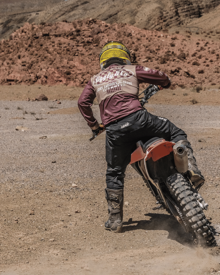 A person riding a motocross bike in the desert in a brown Uggly&Co MX shirt