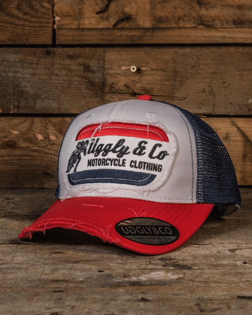 A red, white and blue flat-track cap with a frayed Uggly&Co badge featuring a motorcycle racing theme, placed on a wooden surface.
