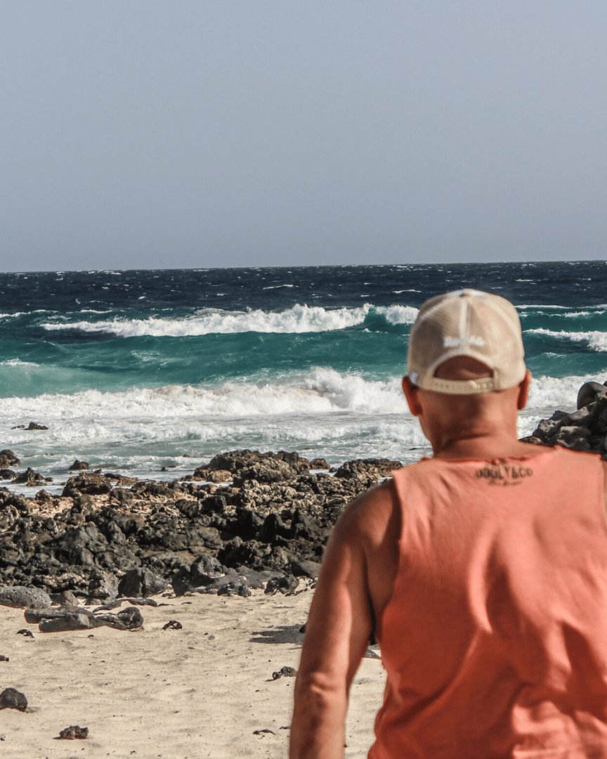 Rear view of a model on a beach wearing a beige and white Uggly & Co motorcycle clothing trucker hat with a mesh back