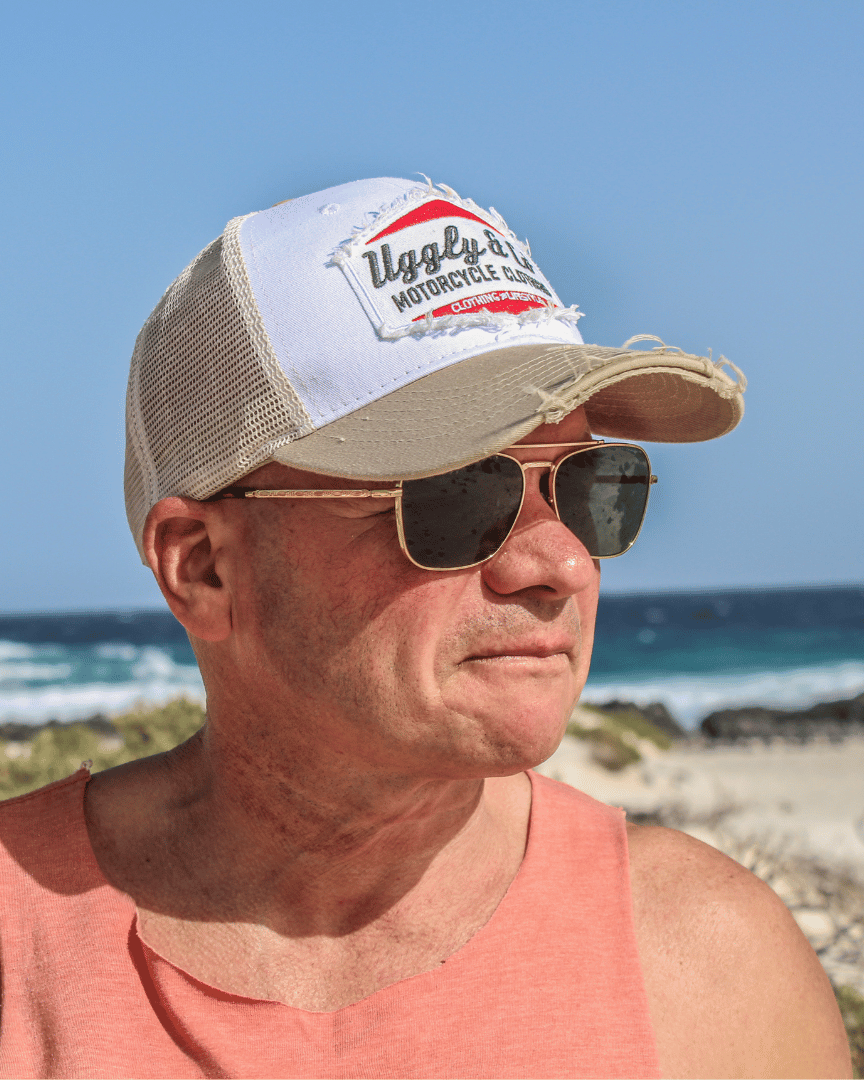 Front view of a model on a beach wearing a beige and white Uggly & Co motorcycle clothing trucker hat with a mesh back and an embroidered logo on the front