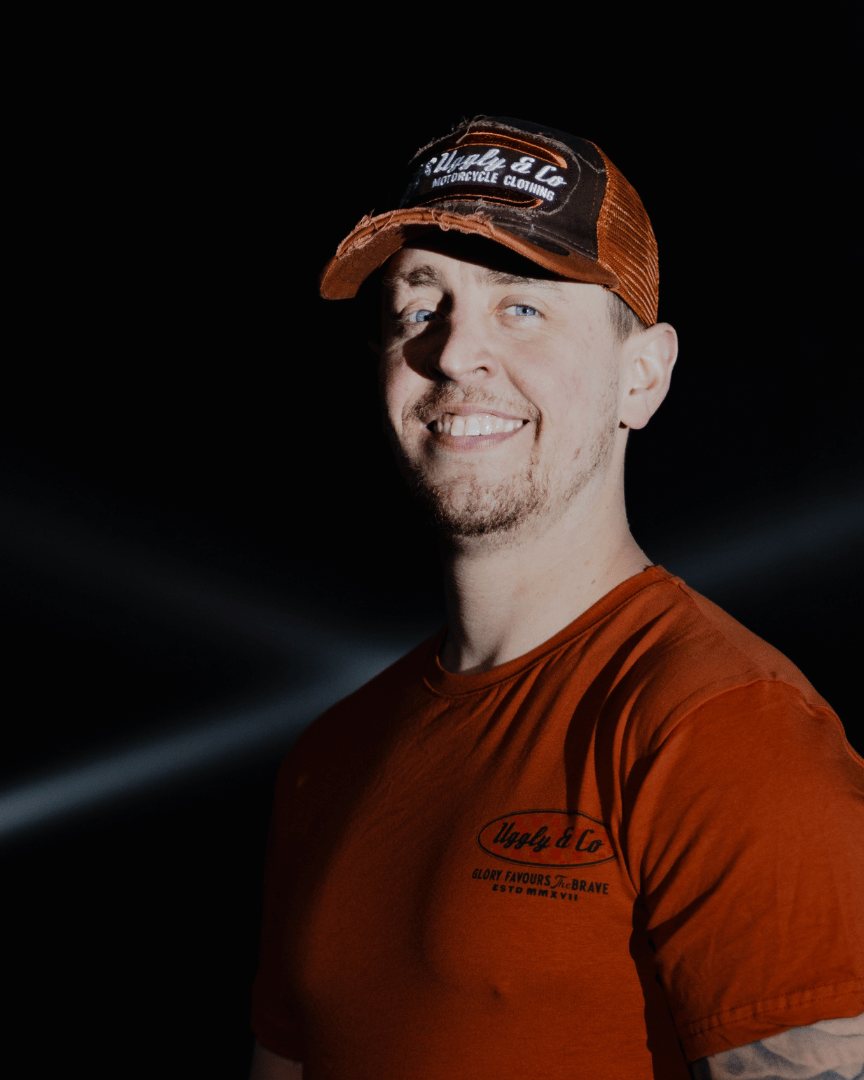 A model smiling while wearing a brown trucker hat with an orange brim, featuring a flat track Uggly&Co logo patch on the front at night