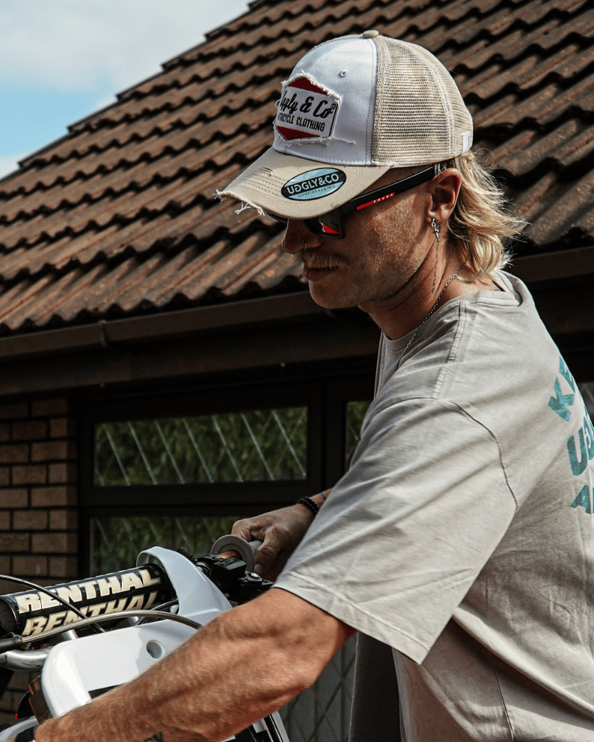 A model holding a motocross bike while wearing a beige and white Uggly & Co motorcycle clothing trucker hat with a mesh back and an embroidered logo on the front