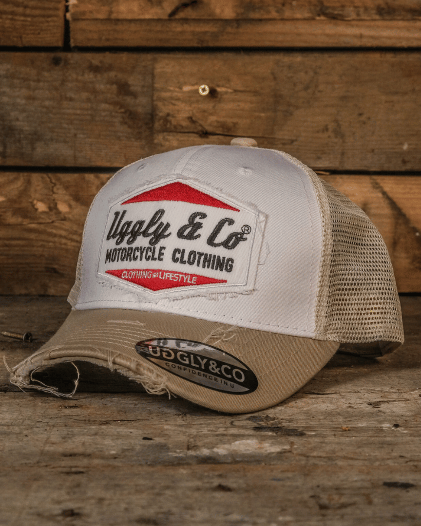 A beige and white Uggly & Co motorcycle clothing trucker hat with a mesh back and an embroidered logo on the front, placed against a wooden background.