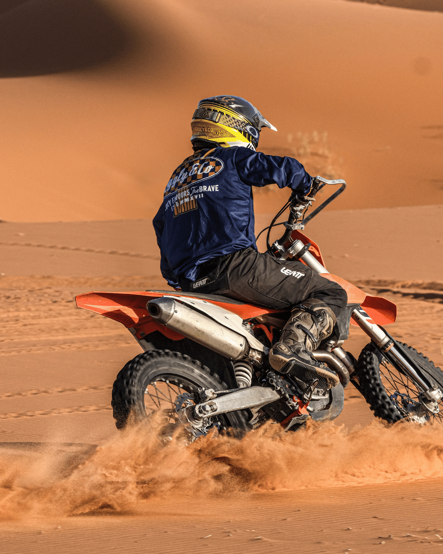 MX rider on a bike in the desert in a blue motocross jersey and the brand name 'Uggly&Co Motorcycle Clothing' on the back