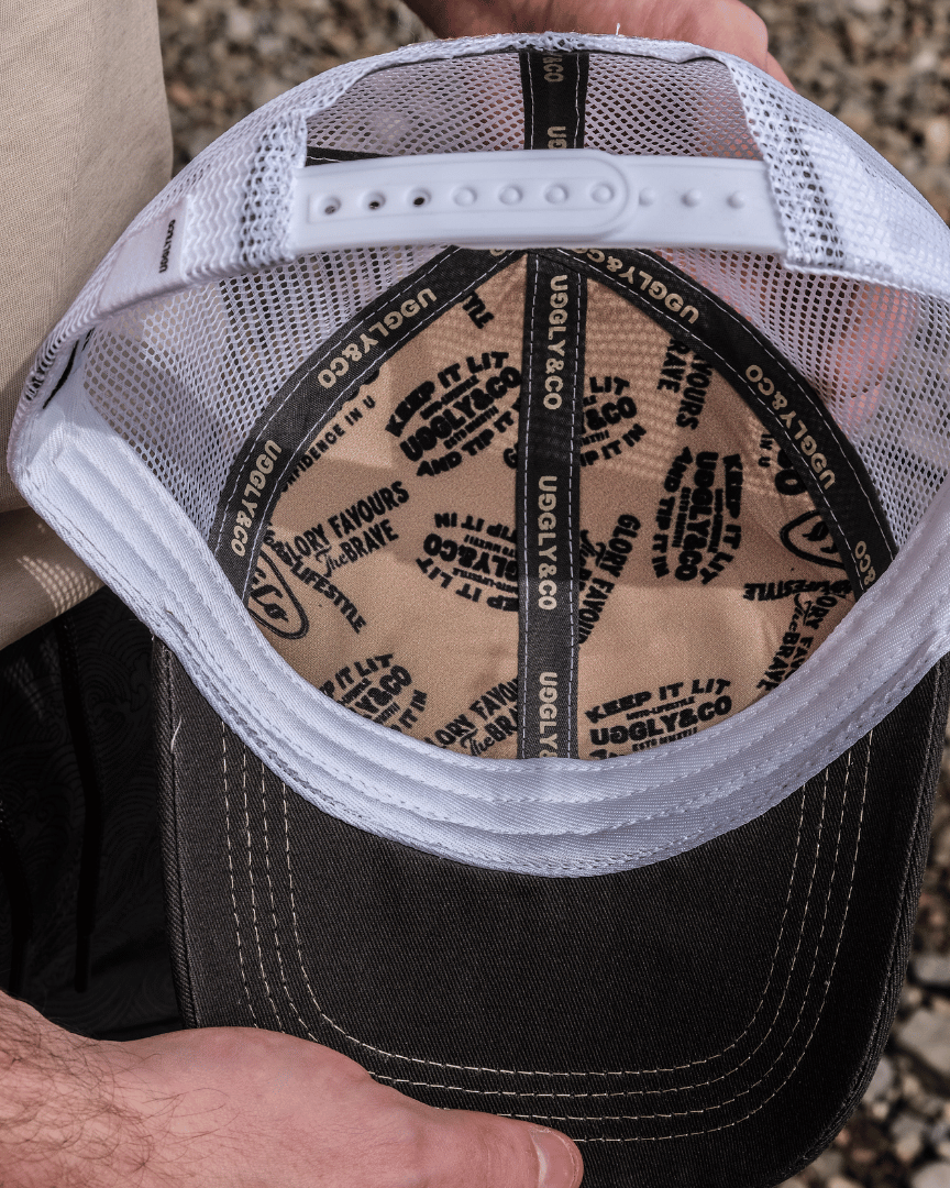 Close up of the inside of a white, beige and brown trucker cap with Uggly&Co branding on the seams with a blurred rocky background.