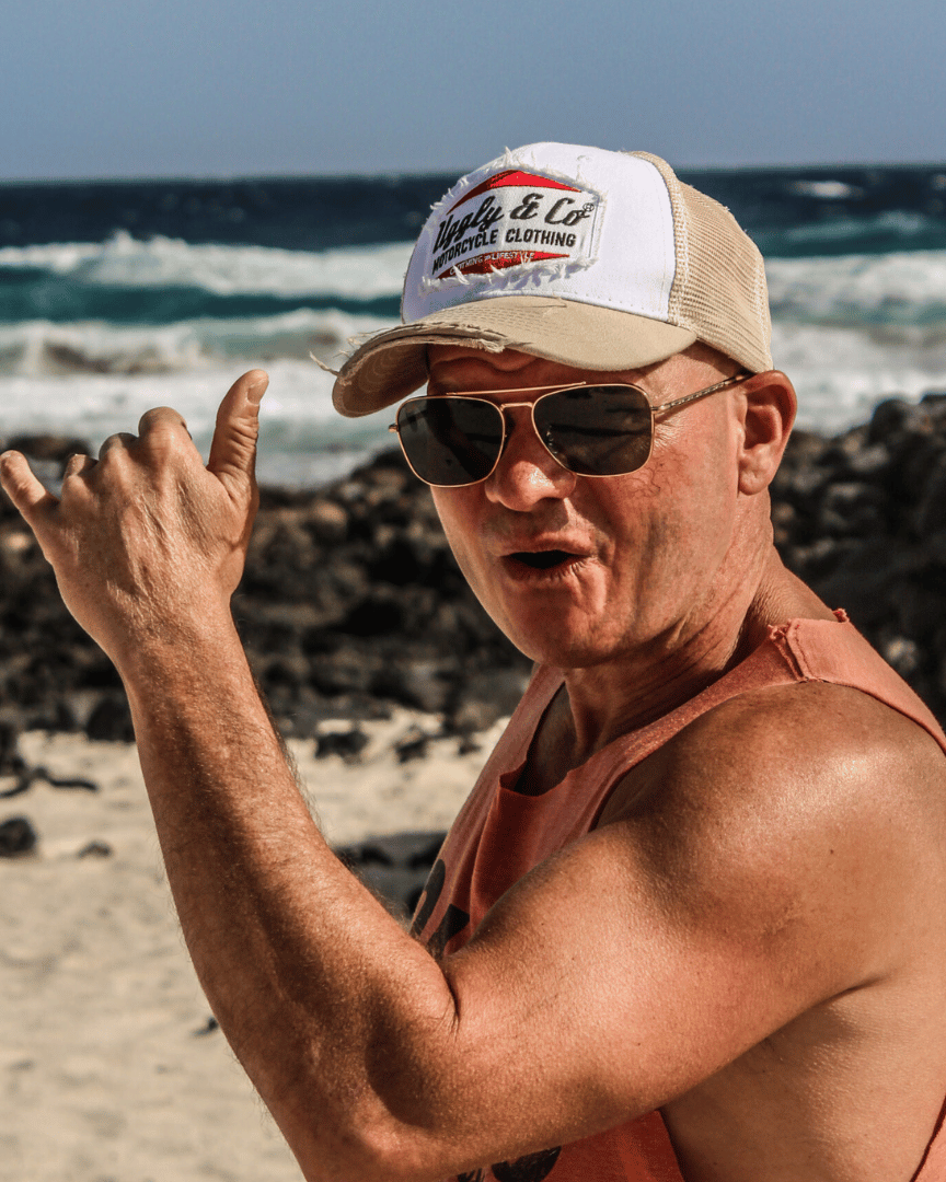 A model on a beach wearing a beige and white Uggly & Co motorcycle clothing trucker hat with a mesh back and an embroidered logo on the front
