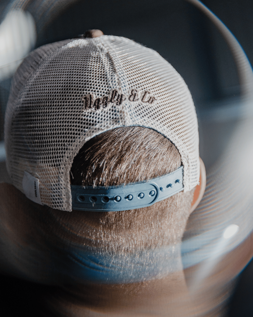 Rear view of a model wearing a dusty blue trucker hat with a brown front panel, featuring a vintage flat track Uggly&Co logo