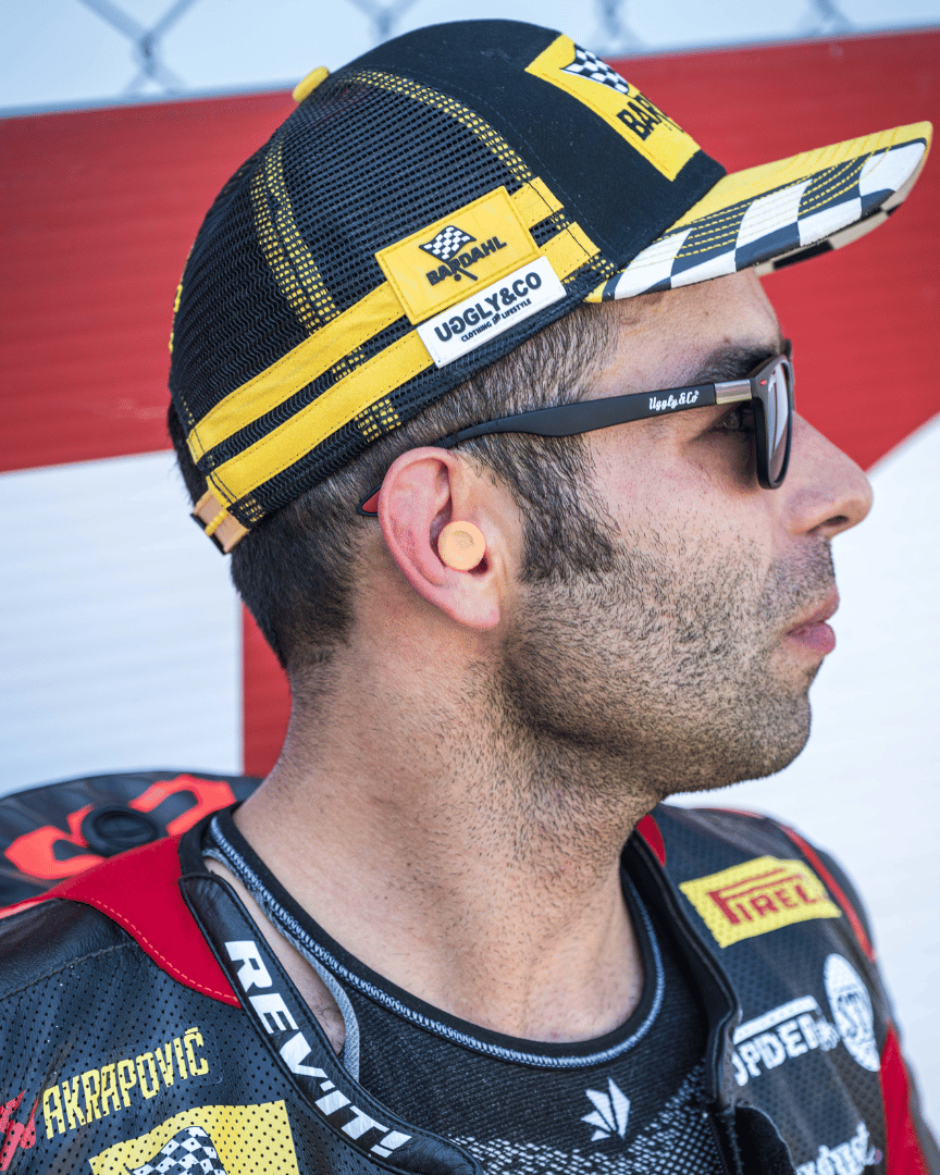 Danilo Pertrucci looking right while wearing a yellow and black Uggly&Co Bardahl Danilo Pertrucci #9 merchandise trucker hat