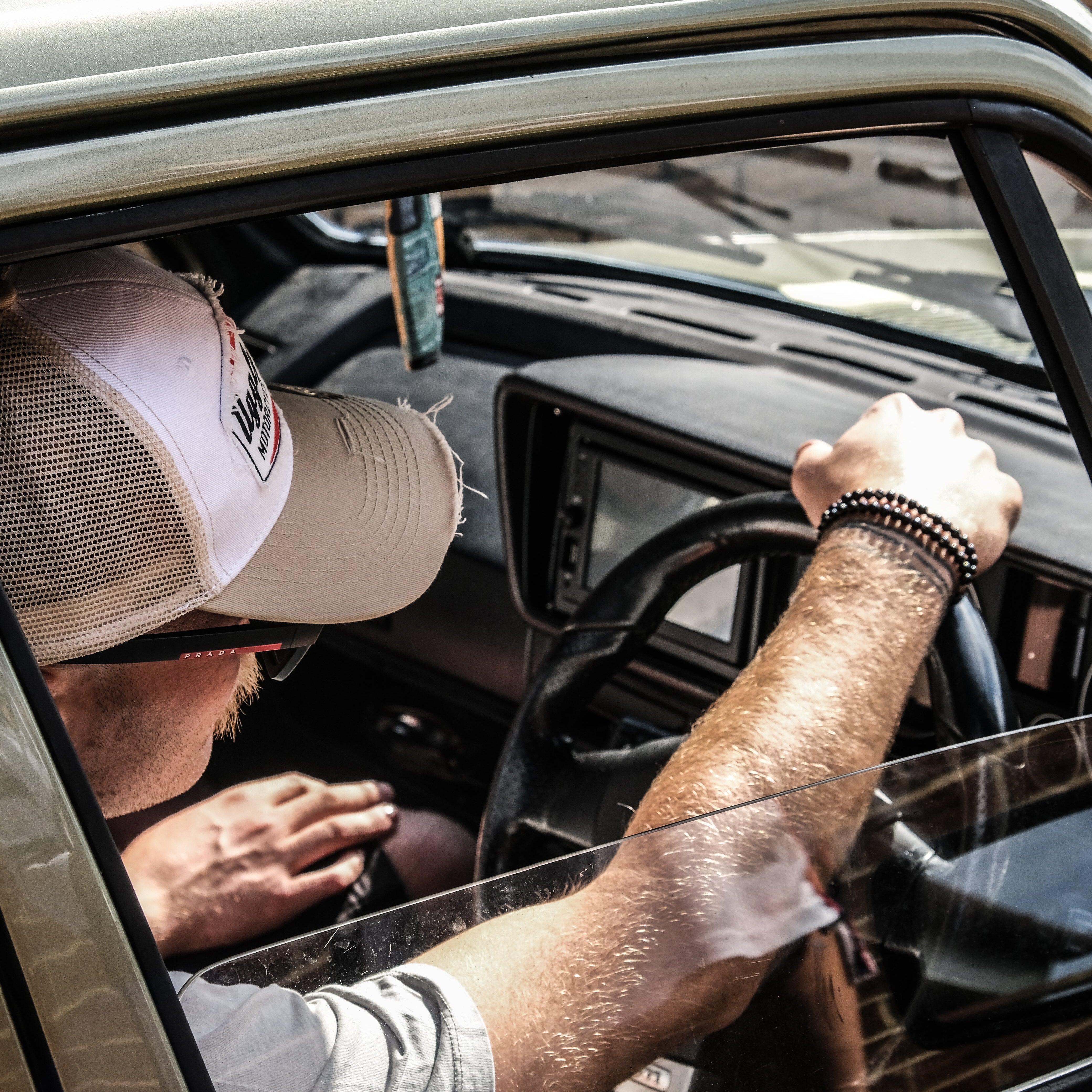 A man wearing a beige trucker hat and light gray t-shirt sits in the driver's seat of a vintage beige car, holding the steering wheel through the open window.