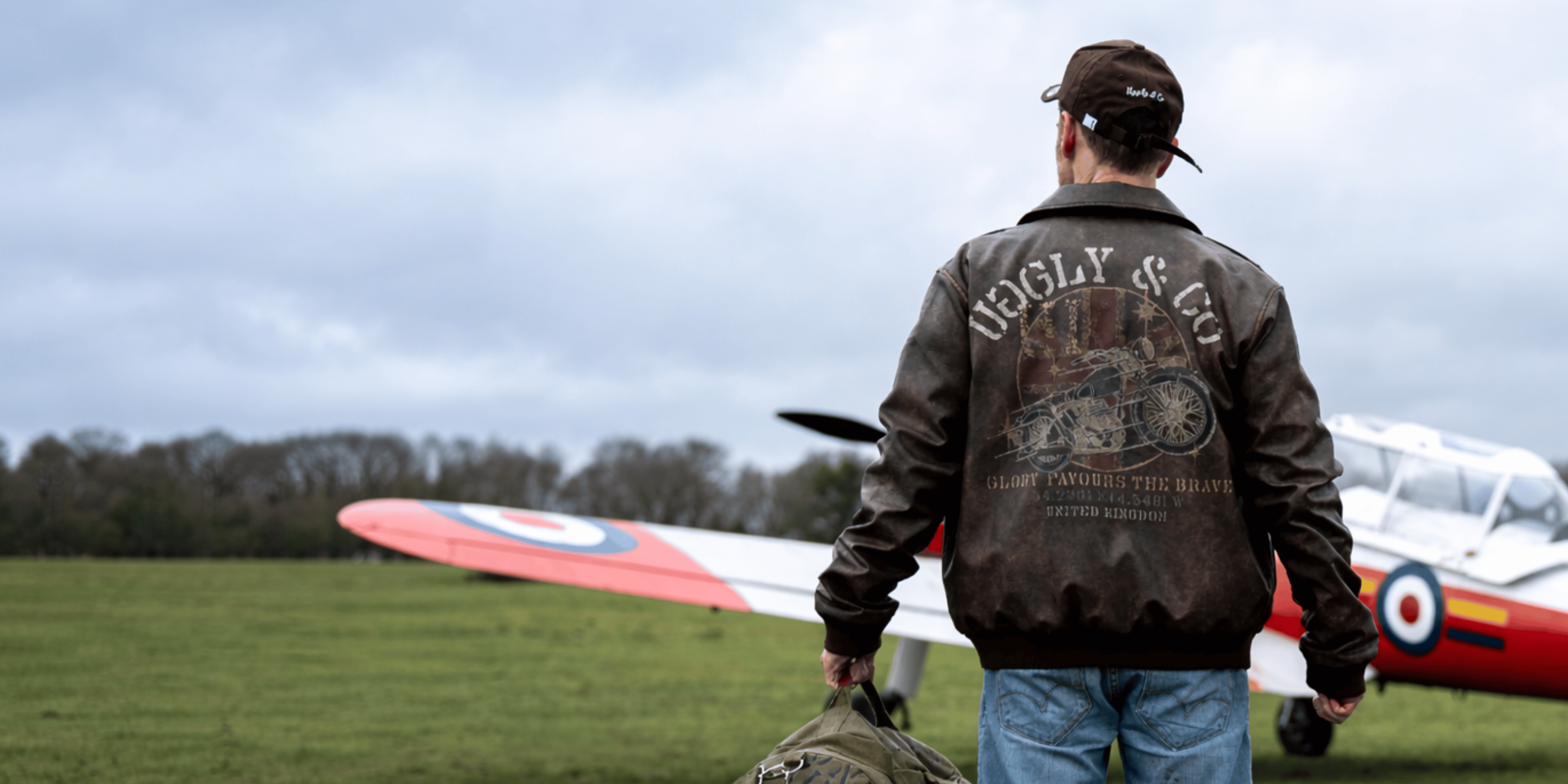A model wearing a brown Uggly&Co aviation jacket on a grass airfield in front of a plane