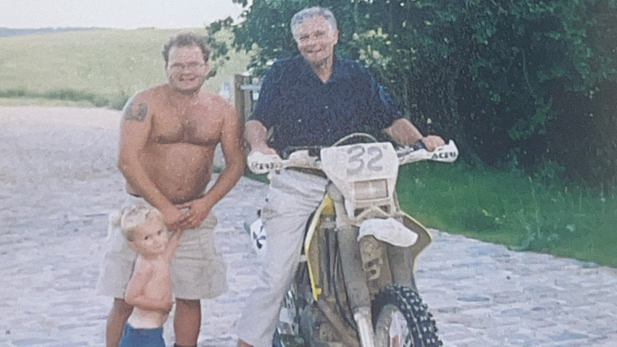 A vintage outdoor photograph showing a shirtless man, an older man sitting on a dirt bike, and a small child standing together on a cobblestone road with trees in the background.