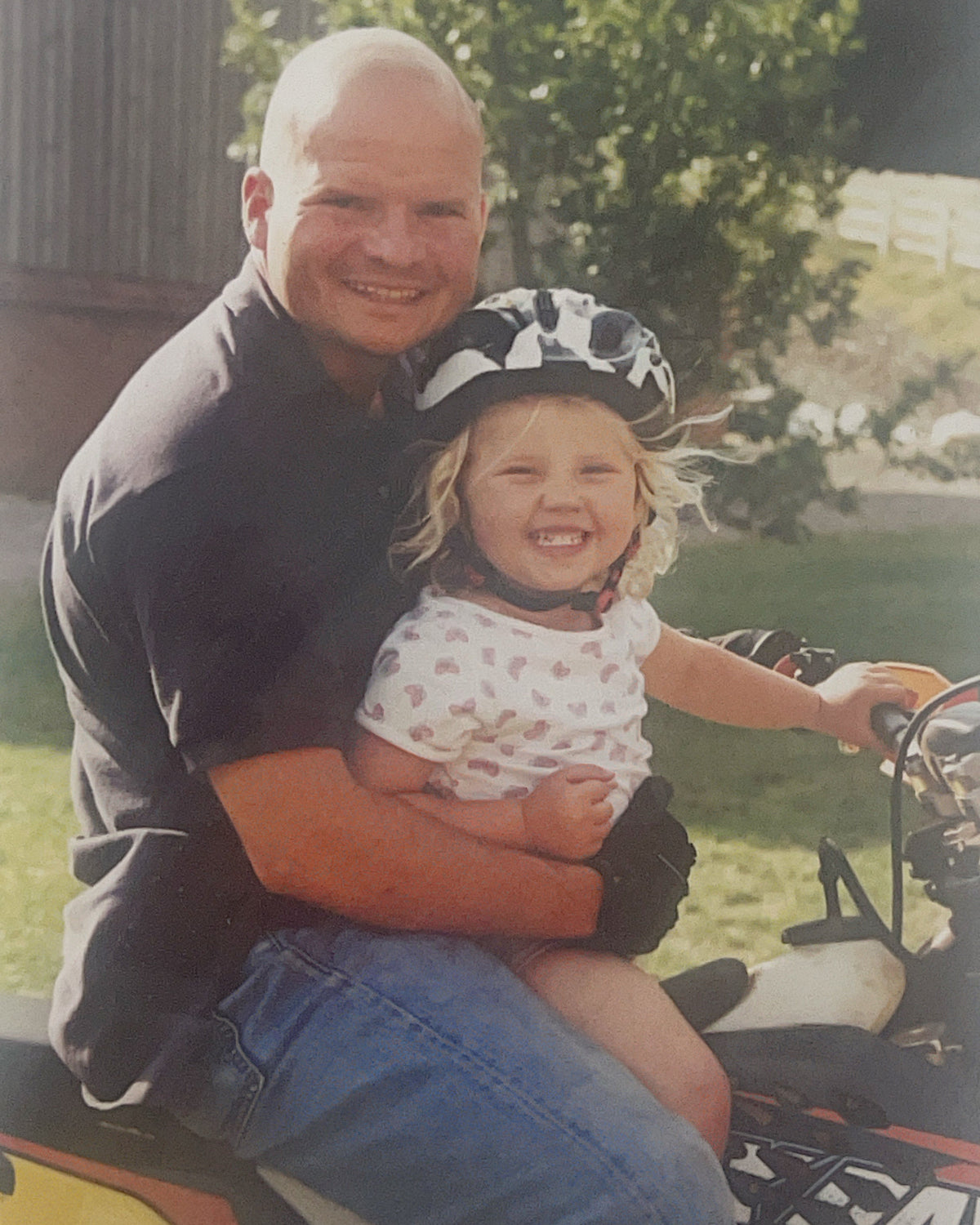 A smiling bald man in a black shirt and blue jeans holds a young child wearing a black-and-white patterned helmet while sitting on a yellow and black ATV outdoors.