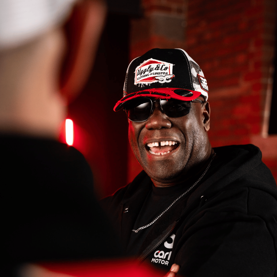 Carl Cox the DJ wearing a cap and sunglasses with a red background