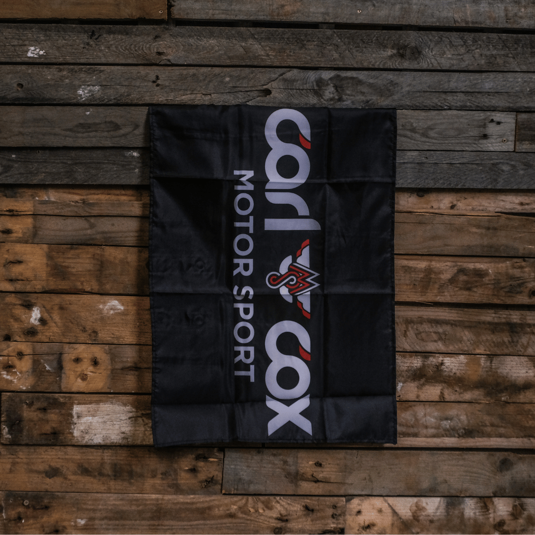 A rectangular black flag with the 'Carl Cox Motorsport' logo in white and red, displayed on a wooden surface.