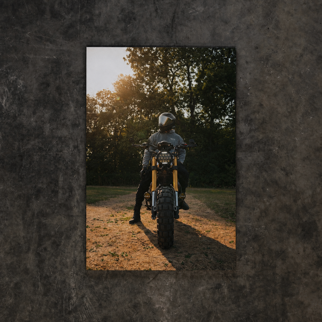 A vertical rectangular wall art print showing a rider on a motorcycle on a dirt path at sunset, centered on a dark textured wall.