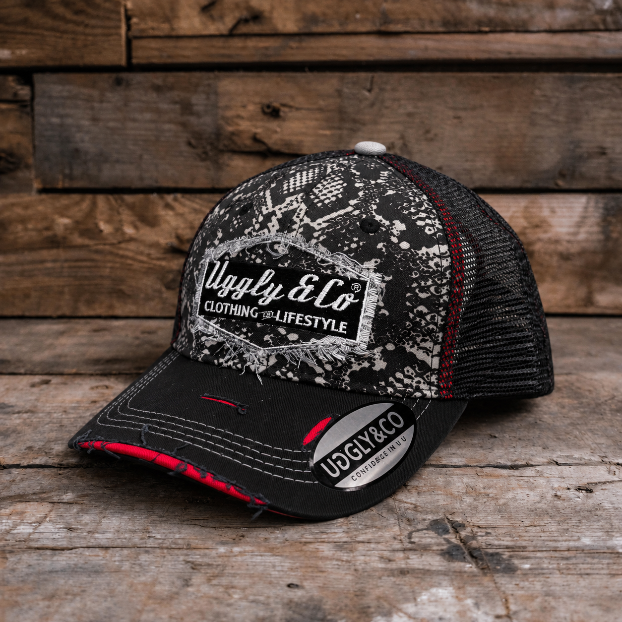 A black and white trucker hat with a distressed pattern on the front panel and brim, featuring a white 'Uggly & Co' logo patch. The back mesh is black with thin red stitching accents, and the hat is photographed on a rustic wooden plank surface.