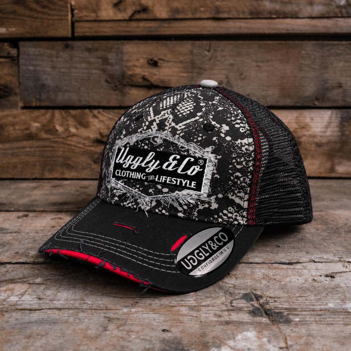 A black and white trucker hat with a distressed pattern on the front panel and brim, featuring a white 'Uggly & Co' logo patch. The back mesh is black with thin red stitching accents, and the hat is photographed on a rustic wooden plank surface.