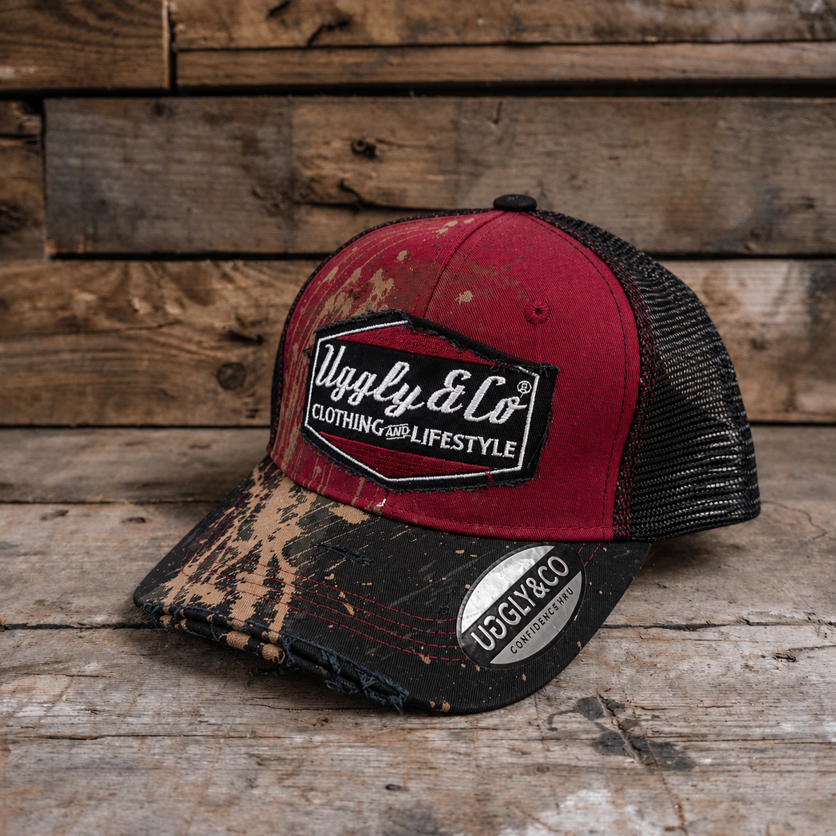 A distressed trucker hat with a red front panel, black mesh back, and a black brim featuring a brown and beige Mud splatter motorcross pattern. The front has a black patch with the text 'Uggly & Co CLOTHING AND LIFESTYLE'.