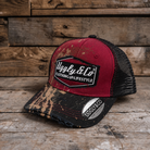 A distressed trucker hat with a red front panel, black mesh back, and a black brim featuring a brown and beige Mud splatter motorcross pattern. The front has a black patch with the text 'Uggly & Co CLOTHING AND LIFESTYLE'.