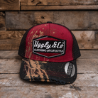 A trucker-style cap with a distressed red front panel, black mesh back, and a camouflage-patterned brim, displayed on a rustic wooden surface.