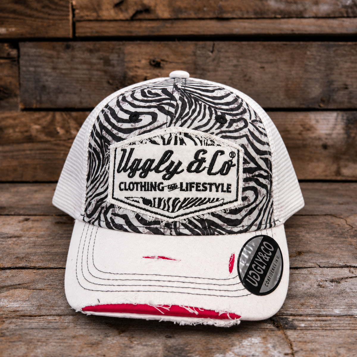 A distressed trucker cap with a black and white zebra print front panel, white mesh back, and a white brim with frayed detailing and a red accent on the underside. The front features a patch for 'Uggly & Co Clothing & Lifestyle' and a circular 'UGGLY & CO' sticker on the brim, set against a rustic wooden plank background.