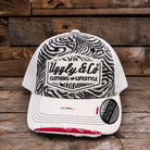 A distressed trucker cap with a black and white zebra print front panel, white mesh back, and a white brim with frayed detailing and a red accent on the underside. The front features a patch for 'Uggly & Co Clothing & Lifestyle' and a circular 'UGGLY & CO' sticker on the brim, set against a rustic wooden plank background.