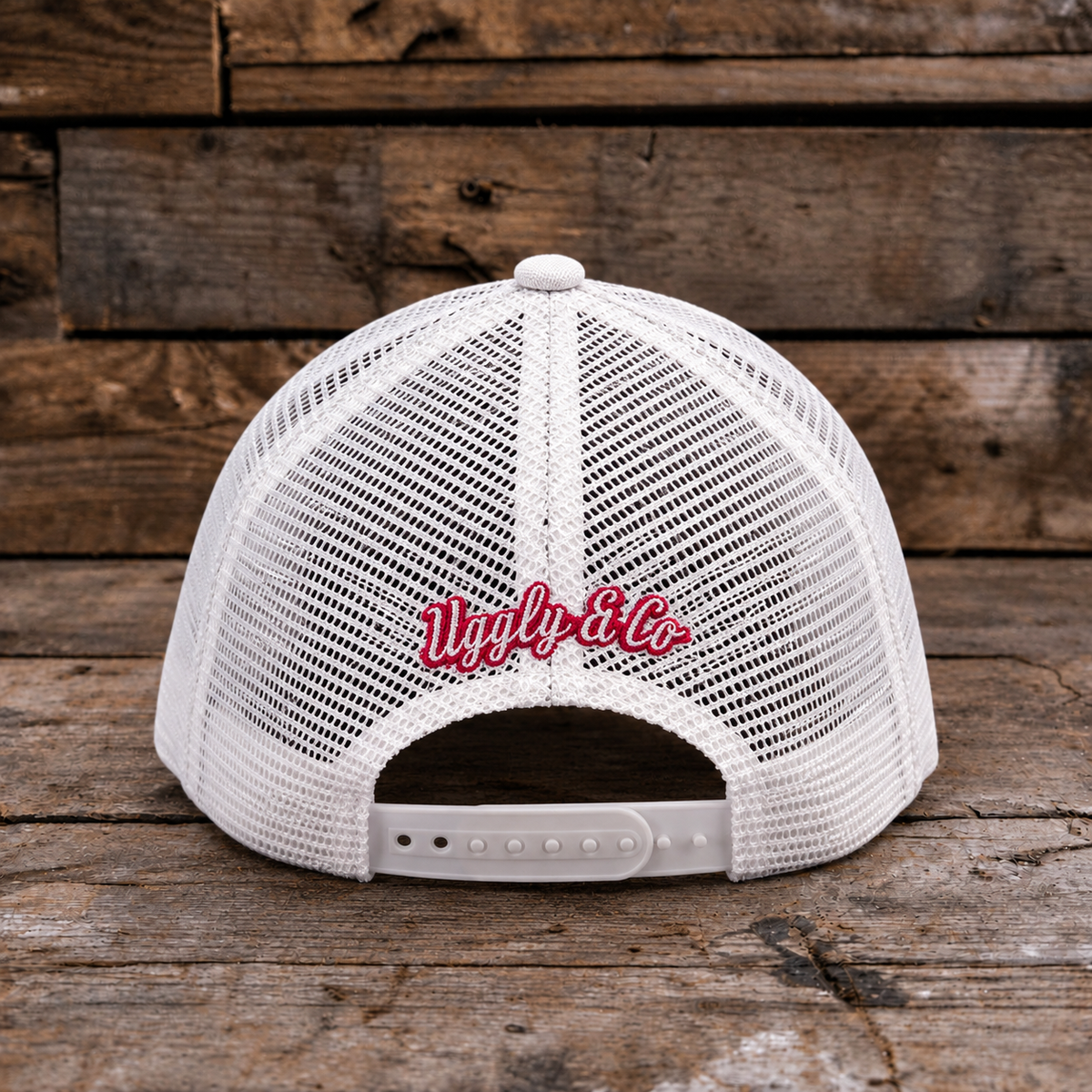 White mesh trucker hat shown from the back, featuring red embroidered cursive text reading 'Ugly & Co.' on the center back, photographed against a rustic wooden plank background.
