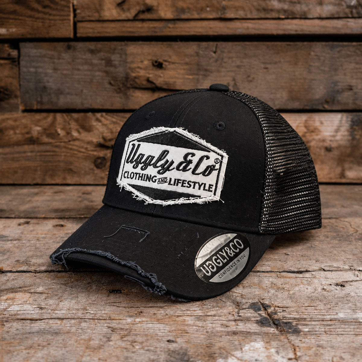 A black trucker-style cap with distressed detailing, a white embroidered 'Uggly & Co' logo on the front, and a matching sticker on the brim, displayed on a rustic wooden surface.