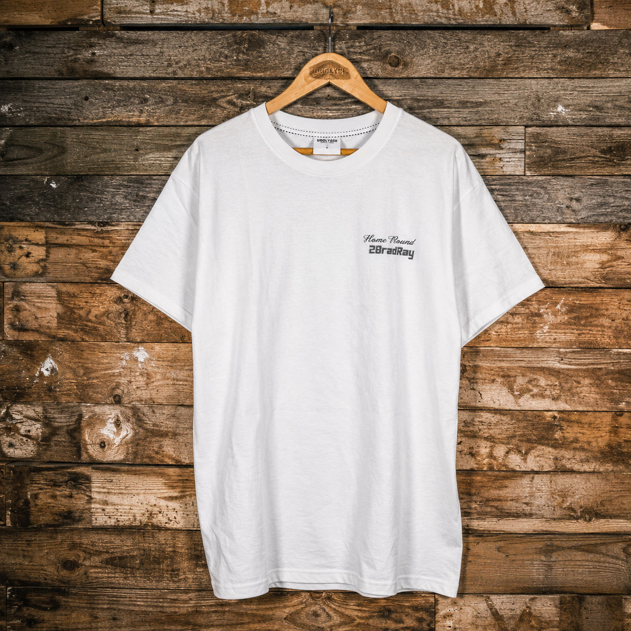 A white short-sleeve crewneck t-shirt with a small black graphic on the left chest, hanging on a wooden hanger against a rustic brown wood plank wall.