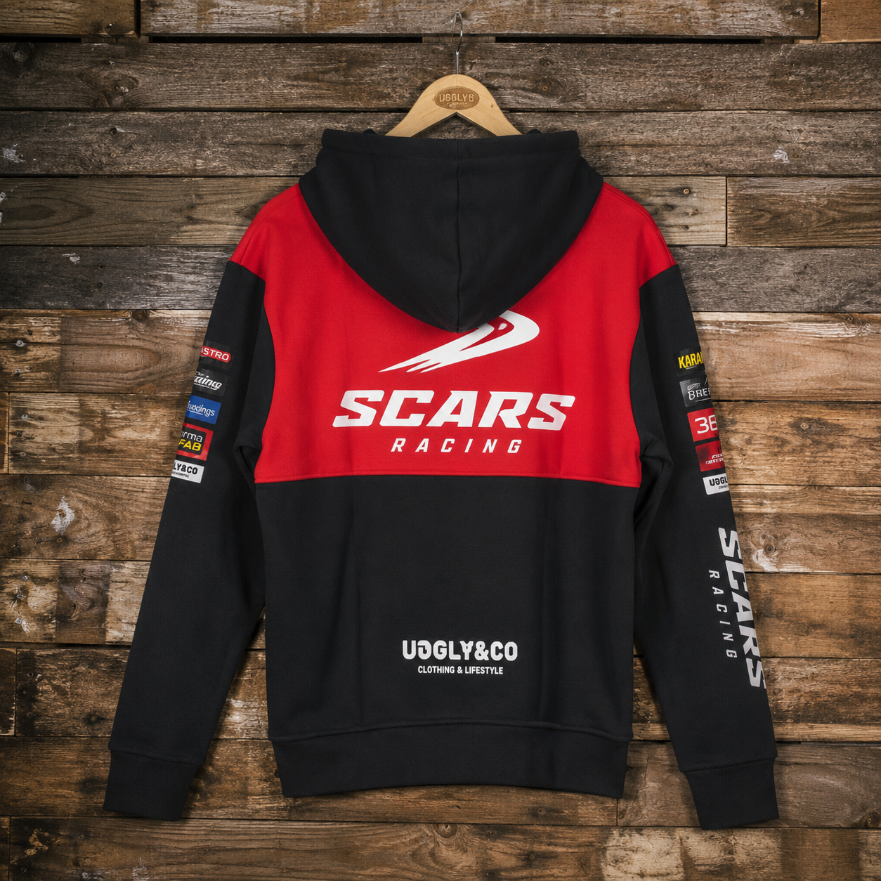 A black and red hooded sweatshirt shown from the back, hanging on a wooden hanger against a rustic wood plank wall. The hoodie features a bold red upper back with a large white 'SCARS RACING' logo, black sleeves, and multiple sponsor-style patches on the sleeves.