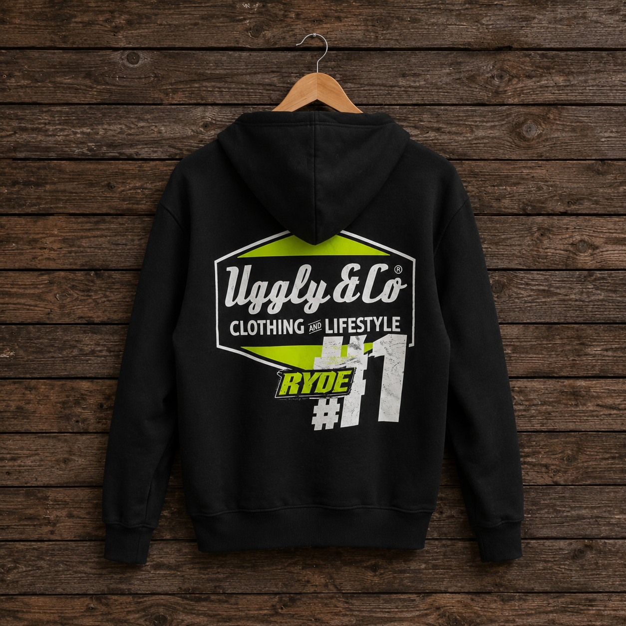 A black hoodie hanging on a wooden hanger against a dark rustic wood plank wall. The back of the hoodie features a large graphic print with the text 'Ugly & Co CLOTHING LIFESTYLE', a lime green hexagon shape, and 'RYDE #11'.