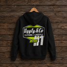 A black hoodie hanging on a wooden hanger against a dark rustic wood plank wall. The back of the hoodie features a large graphic print with the text 'Ugly & Co CLOTHING LIFESTYLE', a lime green hexagon shape, and 'RYDE #11'.