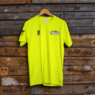 A bright neon yellow t-shirt hanging on a wooden hanger against a rustic dark wood plank wall. The shirt features a black graphic '1' on the left chest, a small mascot logo on the right sleeve, and 'Youth' in white script with a black outline across the center chest.