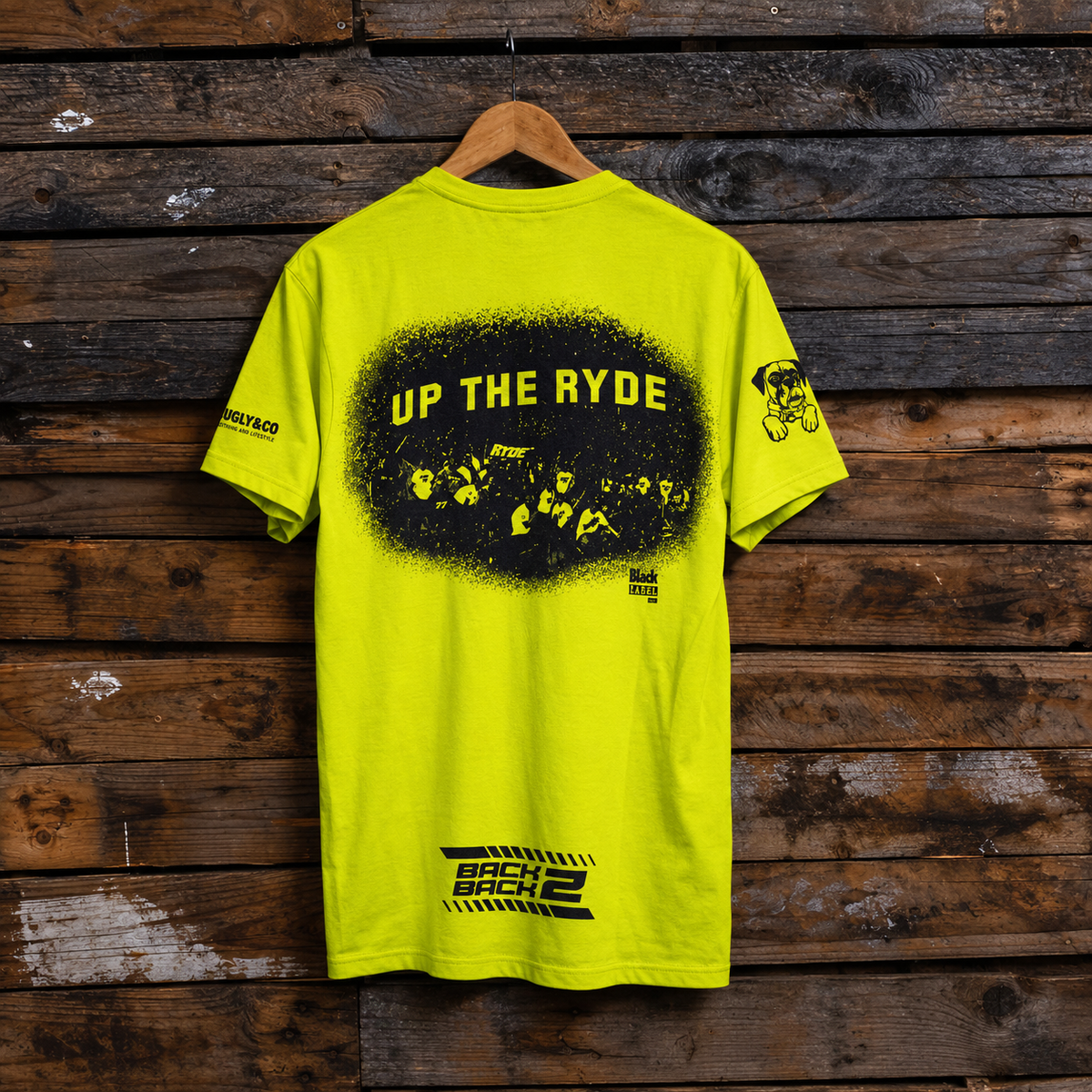 A bright neon yellow t-shirt hanging on a wooden hanger against a rustic dark wood plank wall. The back of the shirt features a large black graphic with yellow text reading 'UP THE RYDE' inside a distressed black circle, a small black graphic on the right sleeve, and a black graphic near the bottom hem.