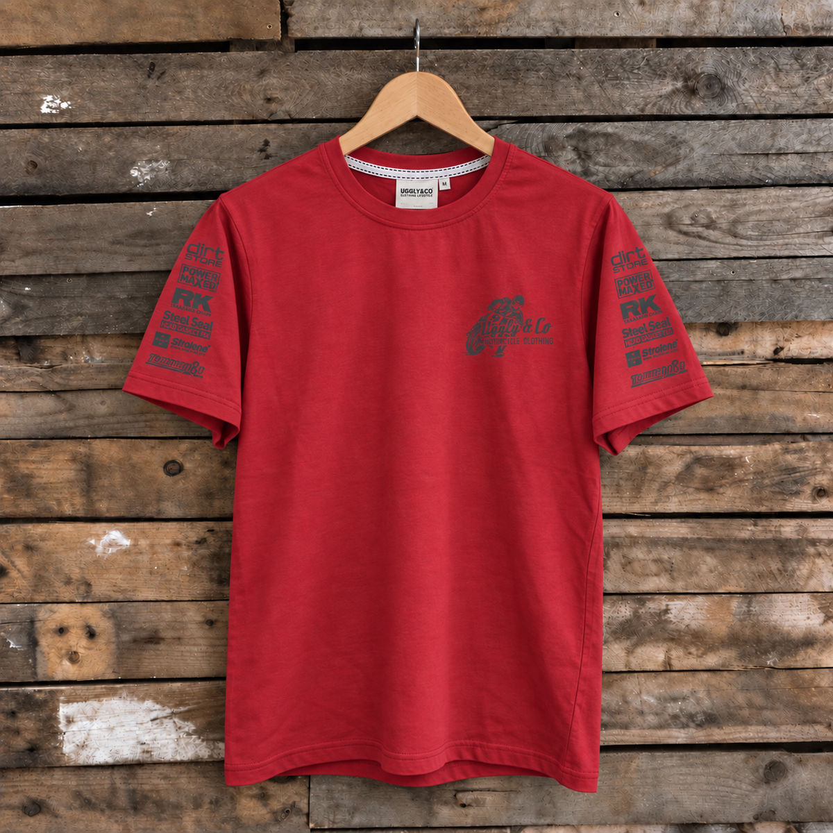 A red short-sleeve t-shirt with dark gray graphic prints on the chest and both sleeves, hanging on a wooden hanger against a rustic wooden plank wall.