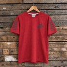 A red short-sleeve t-shirt with dark gray graphic prints on the chest and both sleeves, hanging on a wooden hanger against a rustic wooden plank wall.