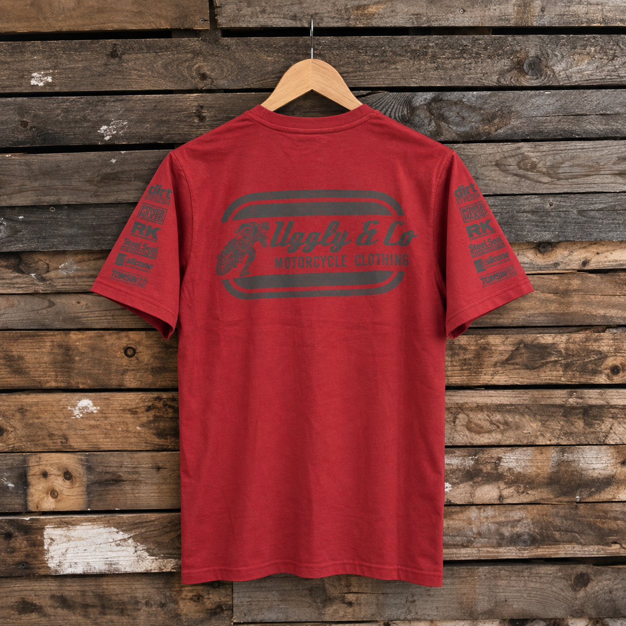 A red short-sleeve t-shirt hanging on a wooden hanger against a rustic wooden plank wall. The back of the shirt features a dark gray graphic with the text 'Riggly & Co Motorcycle Clothing' and 'MFG No.