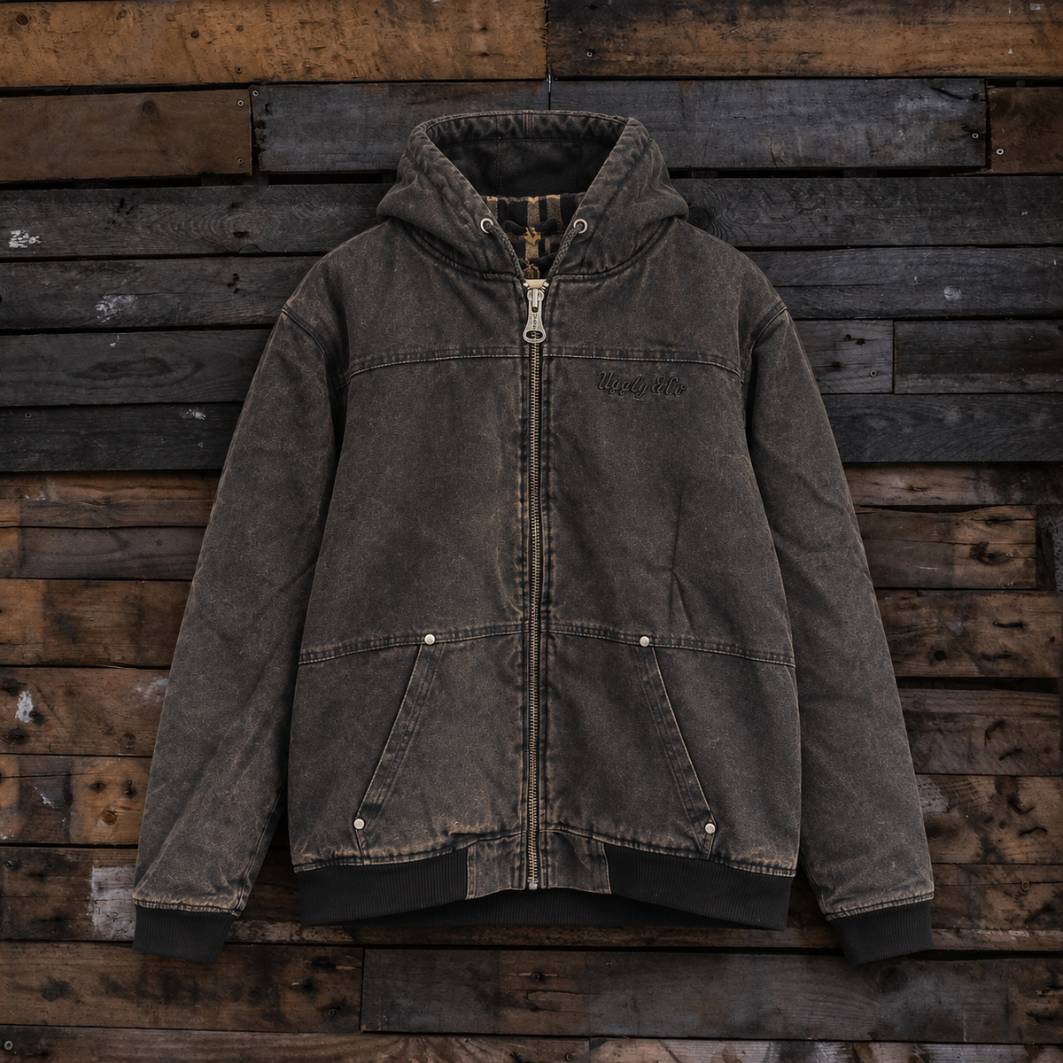 A dark gray washed canvas hooded jacket with a front zipper, displayed flat against a rustic wooden plank background.