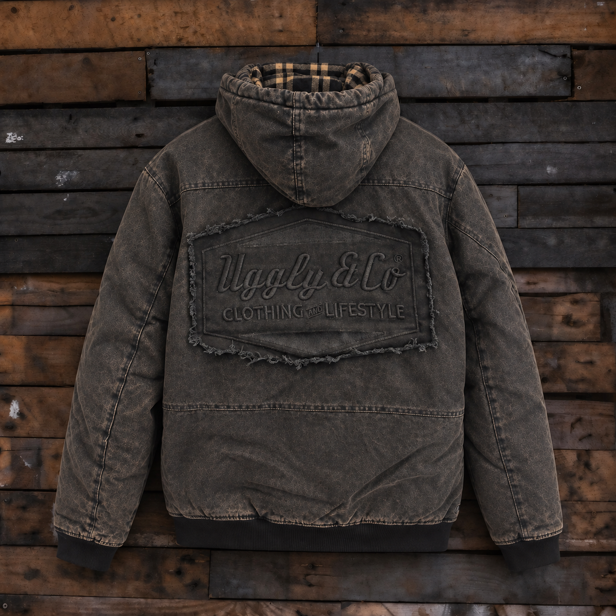 A washed dark brown hooded puffer jacket with a large distressed patch on the back reading 'Uggly & Co Clothing Lifestyle,' displayed against a rustic wooden plank background.