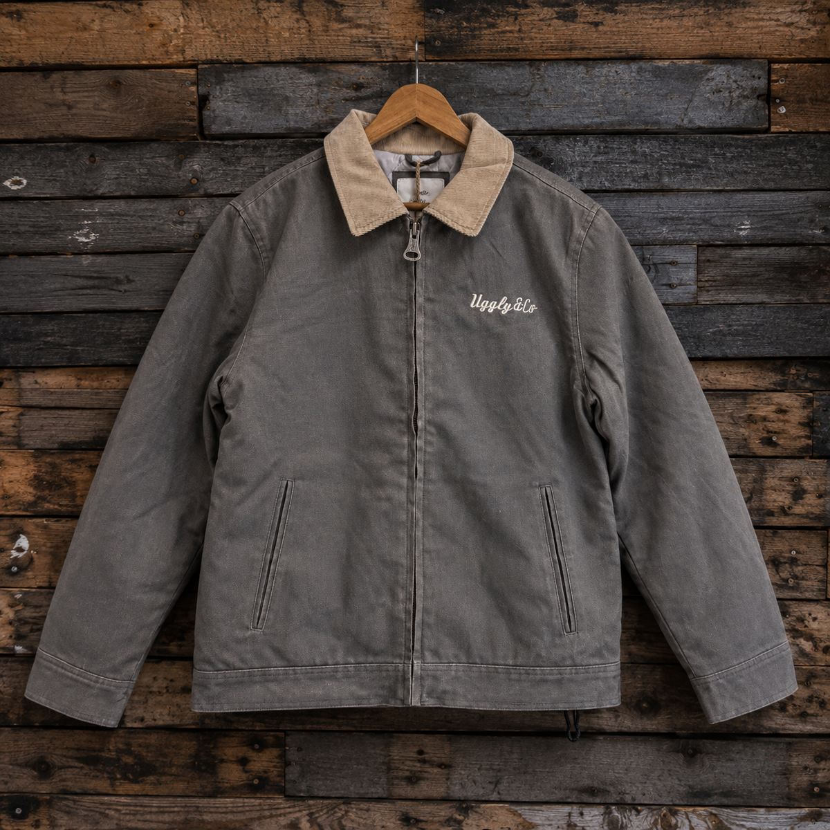 A gray canvas utility jacket with a tan corduroy collar, hanging on a wooden hanger against a rustic dark wood plank wall. The jacket features a front zipper, zippered side pockets, and white embroidered script on the left chest.